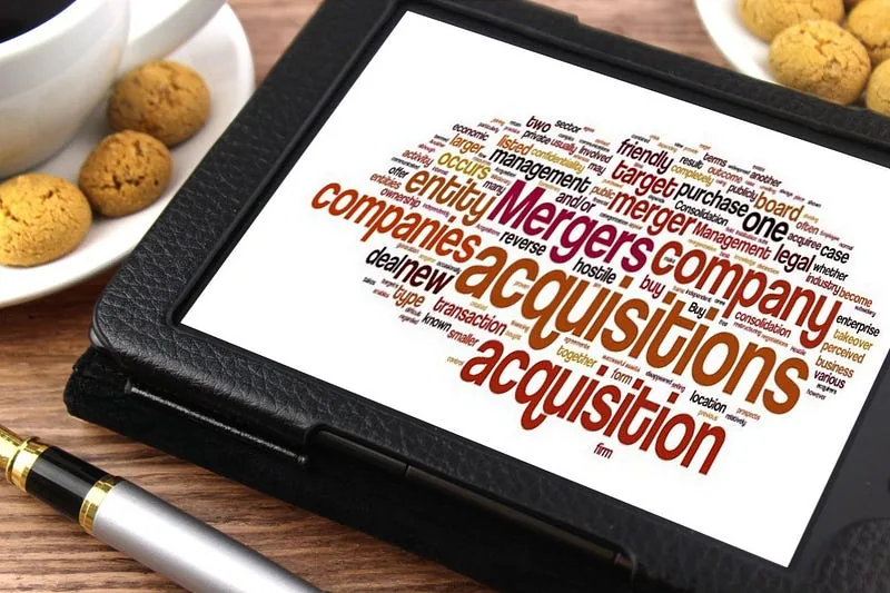 Tablet on a desk displaying a word cloud of merger and acquisition terms, beside coffee and cookies
